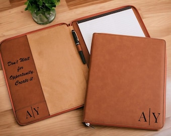May include: A brown leather portfolio with a zipper closure. The portfolio has a pen holder and a notepad. The front of the portfolio has the text "Don't Wait for Opportunity, Create it" and the initials "A|Y" engraved.