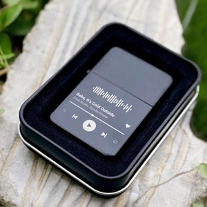 May include: Black metal lighter with a white design featuring a song title, artist, and a play button. The song title is "Baby, It's Cold Outside" by  "Frank Sinatra,  Dorothy Kirsten".