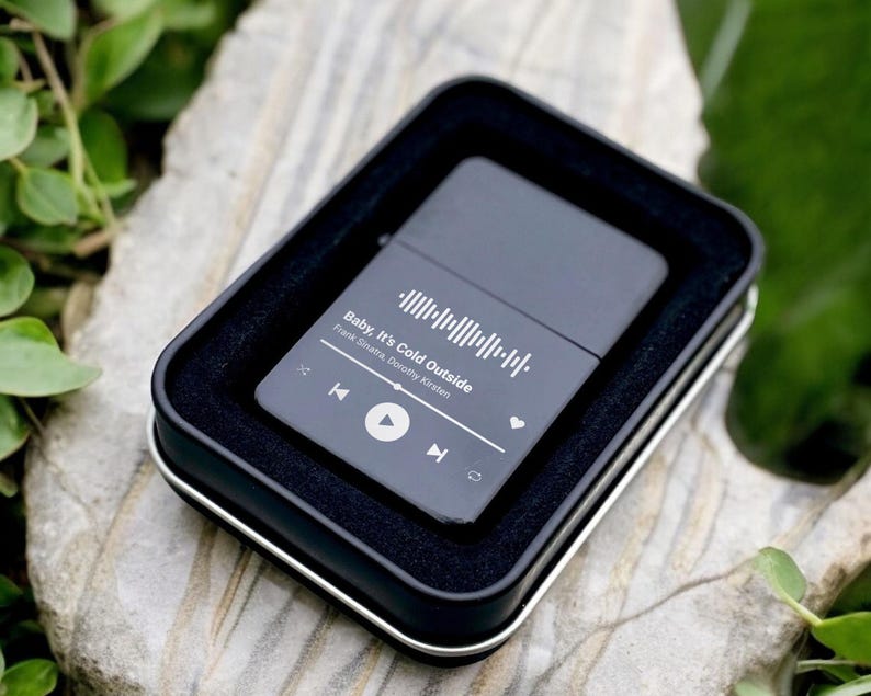 May include: Black metal lighter with a white design featuring a song title, artist, and a play button. The song title is "Baby, It's Cold Outside" by  "Frank Sinatra,  Dorothy Kirsten".