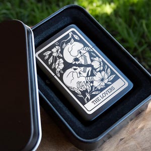 May include: A black metal lighter case with a black and white image of the Lovers tarot card. The card features two skeletons embracing with flowers and vines around them. The text "THE LOVERS" is printed below the image.