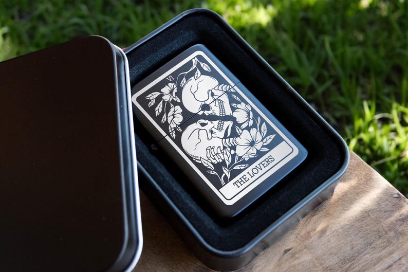 May include: A black metal lighter case with a black and white image of the Lovers tarot card. The card features two skeletons embracing with flowers and vines around them. The text "THE LOVERS" is printed below the image.