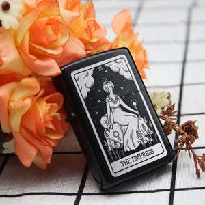 May include: Black lighter with a white design of the Empress tarot card. The card features a woman sitting on a throne with a crown and a symbol of the Empress.