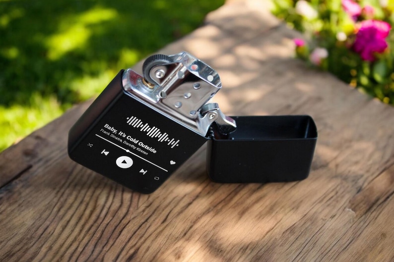 May include: A black lighter with a silver top and a white label that says "Baby, It's Cold Outside" with the song's artists, Frank Sinatra and Dorothy Kirsten, listed below. The label also includes a play button and a barcode.