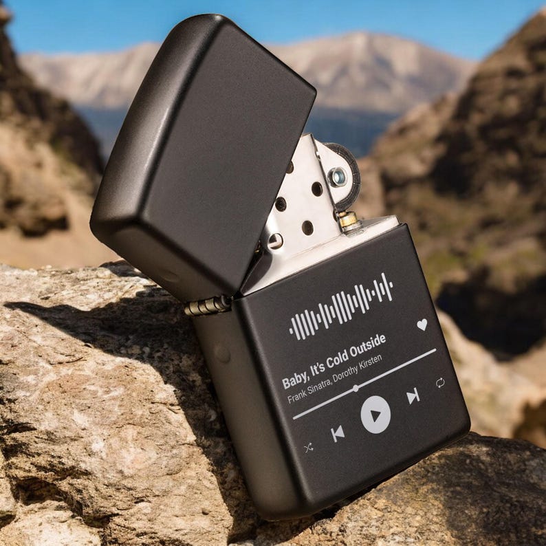 May include: Black lighter with a custom design featuring a music player interface with the song "Baby, It's Cold Outside" by Frank Sinatra and Dorothy Kirsten. The lighter has a heart icon and play, pause, and skip buttons.
