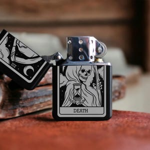 May include: A black lighter with a silver top and a design of the Death tarot card featuring a skeletal figure holding an hourglass. The card reads "DEATH".