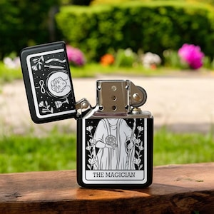 May include: Black lighter with a silver top and a white design of the Magician tarot card. The card features a person holding a wand and a cup, with the text "The Magician" at the bottom.