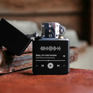 May include: Black lighter with a custom engraved design featuring a song title, artist, and a heart icon. The song title is "Baby, It's Cold Outside" by Frank Sinatra and Dorothy Kirsten. The lighter has a black and silver finish and a built-in music player.