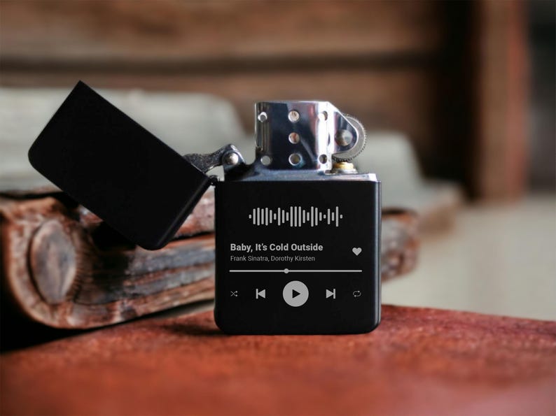 May include: Black lighter with a custom engraved design featuring a song title, artist, and a heart icon. The song title is "Baby, It's Cold Outside" by Frank Sinatra and Dorothy Kirsten. The lighter has a black and silver finish and a built-in music player.