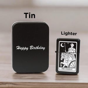 May include: Black tin with the text "Happy Birthday" on it. Next to it is a black lighter with a white design of a tarot card, The Chariot.