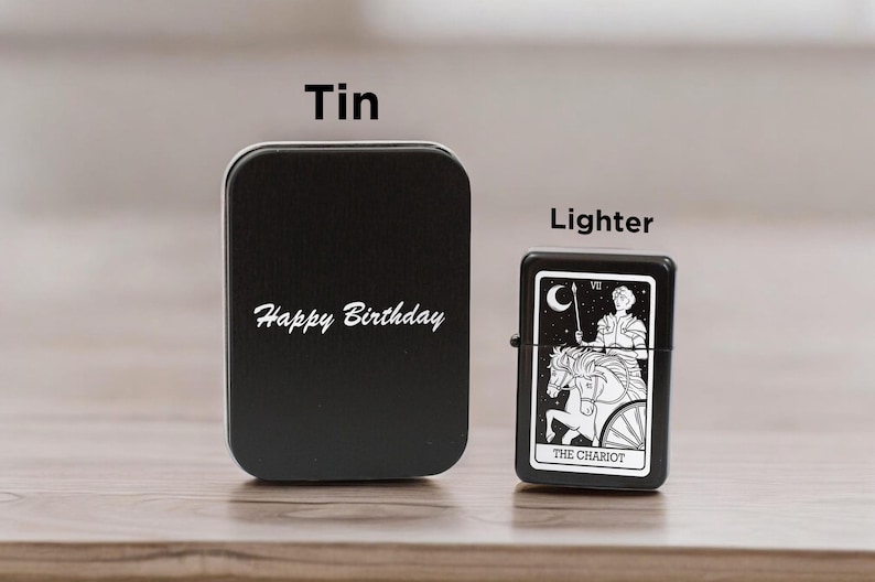 May include: Black tin with the text "Happy Birthday" on it. Next to it is a black lighter with a white design of a tarot card, The Chariot.