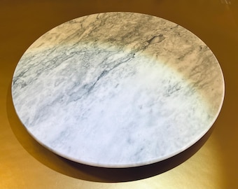 May include: A round marble serving board with a white and gray veined pattern. The board has a smooth surface and is set against a gold-colored background. Ideal for serving appetizers or as a decorative piece.