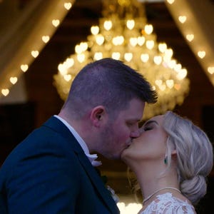 May include: A bride and groom kiss under a canopy of white fabric and string lights shaped like hearts. The lights are strung in a triangular pattern.