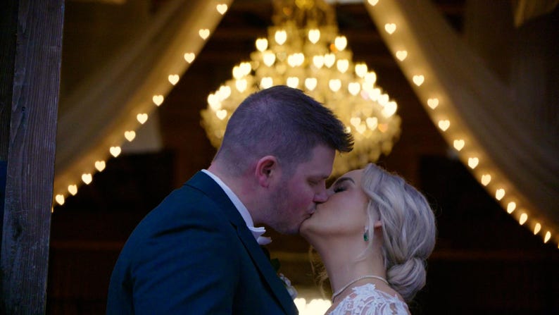 May include: A bride and groom kiss under a canopy of white fabric and string lights shaped like hearts. The lights are strung in a triangular pattern.