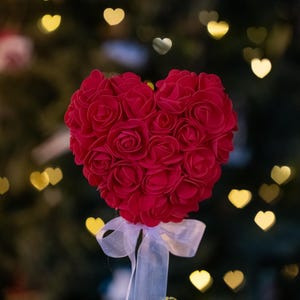 May include: A red heart-shaped arrangement of artificial roses, tied with a white ribbon, against a blurred background of a Christmas tree with golden lights.