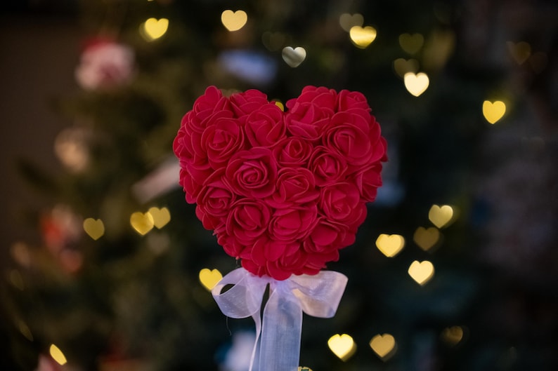 May include: A red heart-shaped arrangement of artificial roses, tied with a white ribbon, against a blurred background of a Christmas tree with golden lights.