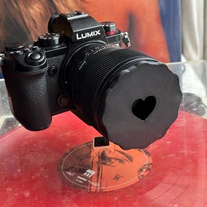 May include: A black camera with a lens cap that has a heart-shaped cutout. The camera is sitting on a red vinyl record.