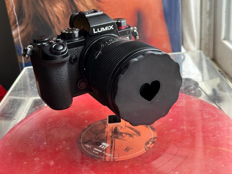May include: A black camera with a lens cap that has a heart-shaped cutout. The camera is sitting on a red vinyl record.