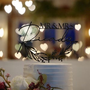 May include: A white wedding cake with a black cake topper that says "Mr. & Mrs. Ricardo" with leafy accents. The cake is in front of a blurred background of white heart-shaped lights.