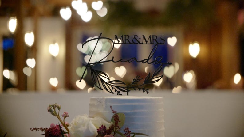 May include: A white wedding cake with a black cake topper that says "Mr. & Mrs. Ricardo" with leafy accents. The cake is in front of a blurred background of white heart-shaped lights.