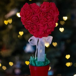 May include: A red heart-shaped rose bush in a red pot with a white ribbon. The bush is surrounded by green foliage and is sitting on a wooden table.
