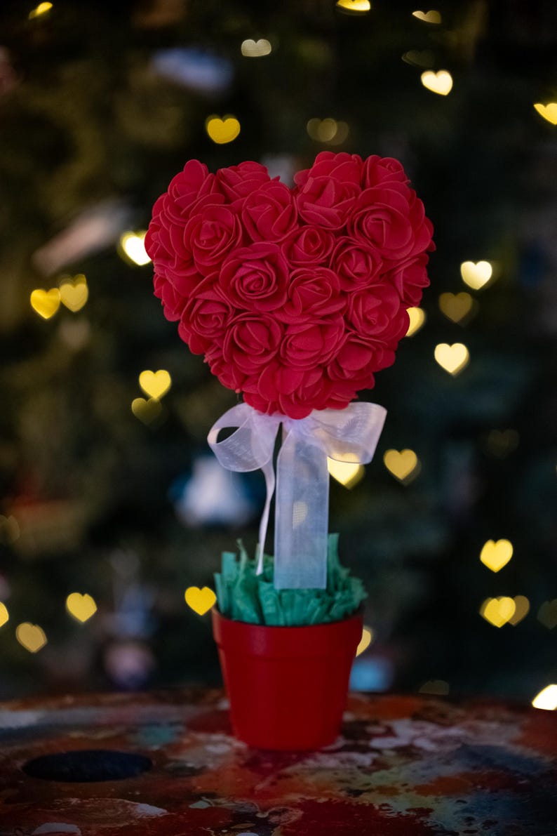 May include: A red heart-shaped rose bush in a red pot with a white ribbon. The bush is surrounded by green foliage and is sitting on a wooden table.