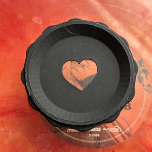 May include: A black, round, 3D printed dish with a heart-shaped cutout. The heart is made of a red and orange material with a marbled pattern.