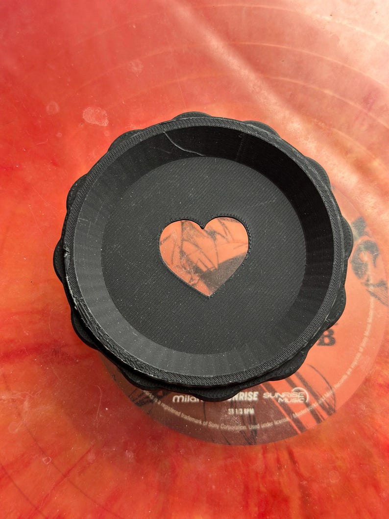 May include: A black, round, 3D printed dish with a heart-shaped cutout. The heart is made of a red and orange material with a marbled pattern.