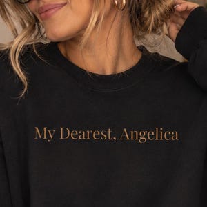 May include: A black long-sleeve crewneck sweatshirt featuring the text My Dearest, Angelica printed in a brown serif font across the center of the chest. The garment has a ribbed neckline, cuffs, and hem, providing a classic fit and style.