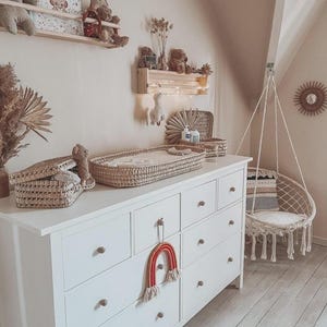 Puede incluir: Una cómoda blanca con pomos de madera y una cesta para cambiar pañales tejida. Un adorno de pared en forma de arcoíris está sujeto a la cómoda. Una silla colgante y objetos decorativos completan la decoración de la guardería.