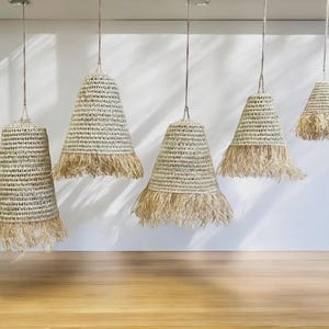 Op de afbeelding: Vijf hanglampen met geweven, klokvormige kappen. De kappen zijn in een natuurlijke beige kleur met een gefranjerde onderrand. De lampen hangen aan dunne koorden aan het plafond, boven een houten vloer.