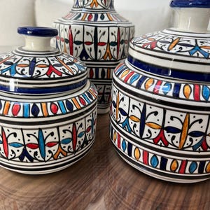 Op de afbeelding: Drie keramische decoratieve stukken met ingewikkelde handgeschilderde ontwerpen. De stukken omvatten een hoge vaas en twee potten met deksel. De ontwerpen hebben een herhalend patroon van zwarte, witte, blauwe, rode en oranje vormen. De stukken staan op een reflecterend oppervlak.