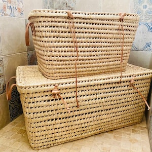 May include: Two rectangular woven storage baskets made from natural straw. They have leather handles and closures. The larger basket is at the bottom, with a smaller one stacked on top. These baskets are suitable for organizing items in a home.