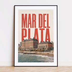 以下が含まれることがあります： 「MAR DEL PLATA」の赤い文字が印象的な、額装されたアートプリント。海岸の風景が描かれており、海辺に建つ2つの壮大な建物が特徴的です。ビーチと水面が前景に描かれています。
