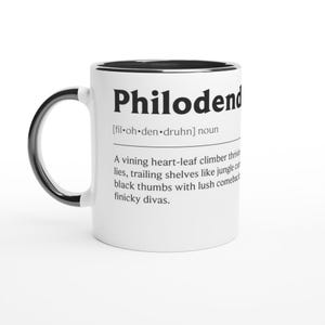 Può includere: Tazza in ceramica bianca con interno e manico neri. La tazza presenta la parola "Philodend" in grassetto nero, insieme a una definizione. La definizione descrive una pianta rampicante.