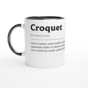 Könnte beinhalten: Weißer Keramikbecher mit schwarzem Henkel und Rand. Der Becher zeigt das Wort "Croquet" in fetten schwarzen Buchstaben sowie eine Definition des Spiels. Ein humorvolles Geschenk für Krocketspieler.