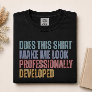 Op de afbeelding: Een zwart t-shirt met ronde hals en de verweerde tekst "DOES THIS SHIRT MAKE ME LOOK PROFESSIONALLY DEVELOPED" in gestapelde regels in lichtblauw, paars, roze en geel. Het t-shirt heeft een Comfort Colors merklabel in de binnenhals.
