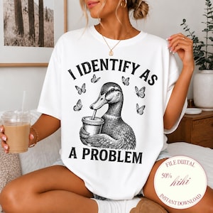 May include: A white short-sleeve t-shirt featuring a black graphic illustration of a duck holding a cup with a straw, surrounded by butterflies. The text "I IDENTIFY AS A PROBLEM" is printed in black capital letters above and below the duck.