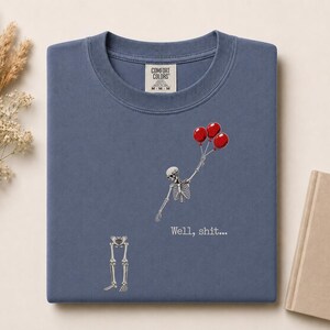 Peut inclure: Un t-shirt bleu &agrave; col rond avec un motif repr&eacute;sentant un squelette tenant trois ballons rouges, le haut du corps s&eacute;par&eacute; des os des jambes. Le texte "Well, shit..." est imprim&eacute; en blanc sous le squelette. Le t-shirt porte une &eacute;tiquette Comfort Colors.