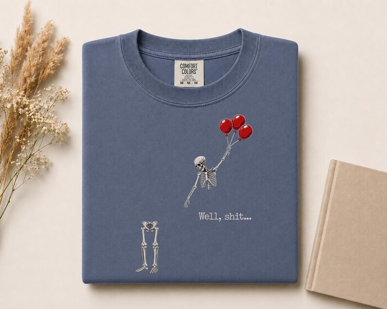 Peut inclure: Un t-shirt bleu &agrave; col rond avec un motif repr&eacute;sentant un squelette tenant trois ballons rouges, le haut du corps s&eacute;par&eacute; des os des jambes. Le texte "Well, shit..." est imprim&eacute; en blanc sous le squelette. Le t-shirt porte une &eacute;tiquette Comfort Colors.