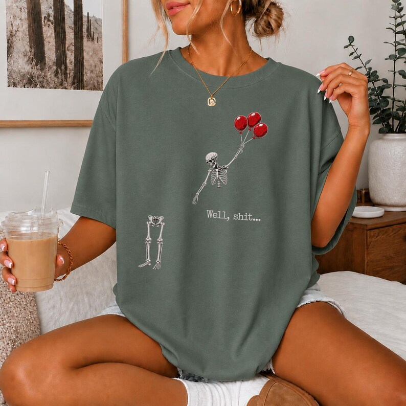 Peut inclure: Un t-shirt vert sauge fonc&eacute; avec un motif repr&eacute;sentant un torse de squelette tenant trois ballons rouges et une section de jambes de squelette s&eacute;par&eacute;e en dessous. Le texte "Well, shit..." est imprim&eacute; en blanc entre les parties du squelette.