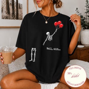 Peut inclure: Un t-shirt noir avec un motif représentant un squelette dont le haut du corps s'envole en tenant trois ballons rouges, tandis que le bassin et les jambes sont séparés en dessous. Le texte "Well, shit..." est imprimé en blanc près du squelette.