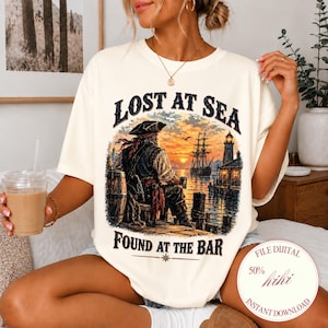 Puede incluir: Una camiseta de manga corta color hueso con un estampado gráfico de un pirata sentado en un muelle al atardecer, con un barco y un faro al fondo. El texto dice LOST AT SEA sobre la imagen y FOUND AT THE BAR debajo de la imagen.