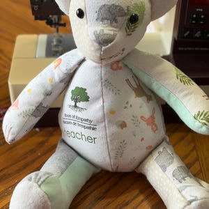 May include: A patchwork teddy bear with a variety of patterned fabrics, including animal and leaf prints. The bear has the text "Roots of Empathy, teacher" printed on its chest. The bear is handmade and sits on a wooden surface.