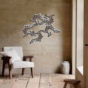 Puede incluir: Una silla blanca con un coj&iacute;n blanco se encuentra en una habitaci&oacute;n con una pared beige. Una gran escultura de pared abstracta hecha de peque&ntilde;os objetos redondos plateados est&aacute; montada en la pared. Una peque&ntilde;a maceta de cer&aacute;mica blanca se encuentra en el suelo junto a un taburete de madera.