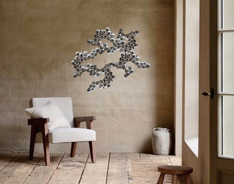 Puede incluir: Una silla blanca con un coj&iacute;n blanco se encuentra en una habitaci&oacute;n con una pared beige. Una gran escultura de pared abstracta hecha de peque&ntilde;os objetos redondos plateados est&aacute; montada en la pared. Una peque&ntilde;a maceta de cer&aacute;mica blanca se encuentra en el suelo junto a un taburete de madera.