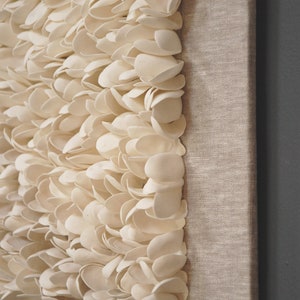 May include: A white wall art piece made of many small, overlapping, white ceramic shapes. The piece is mounted on a canvas with a linen texture.