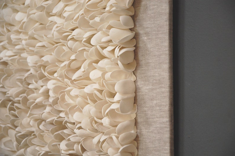 May include: A white wall art piece made of many small, overlapping, white ceramic shapes. The piece is mounted on a canvas with a linen texture.