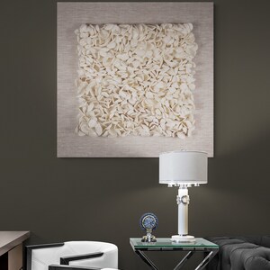 May include: A white, textured wall art piece with a beige frame hangs above a white side table with a glass top. A white lamp with a silver base sits on the table, and a small clock with a blue face is next to it. Two white chairs with black accents are in the foreground.