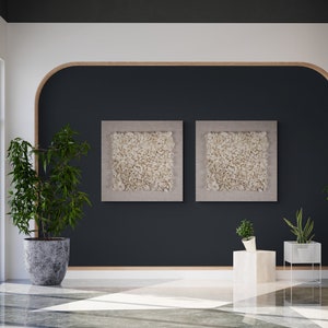 May include: Two framed abstract art prints with a white textured design, hanging on a dark blue wall with a curved wooden frame. The wall is in a modern living room with a potted plant and a side table.