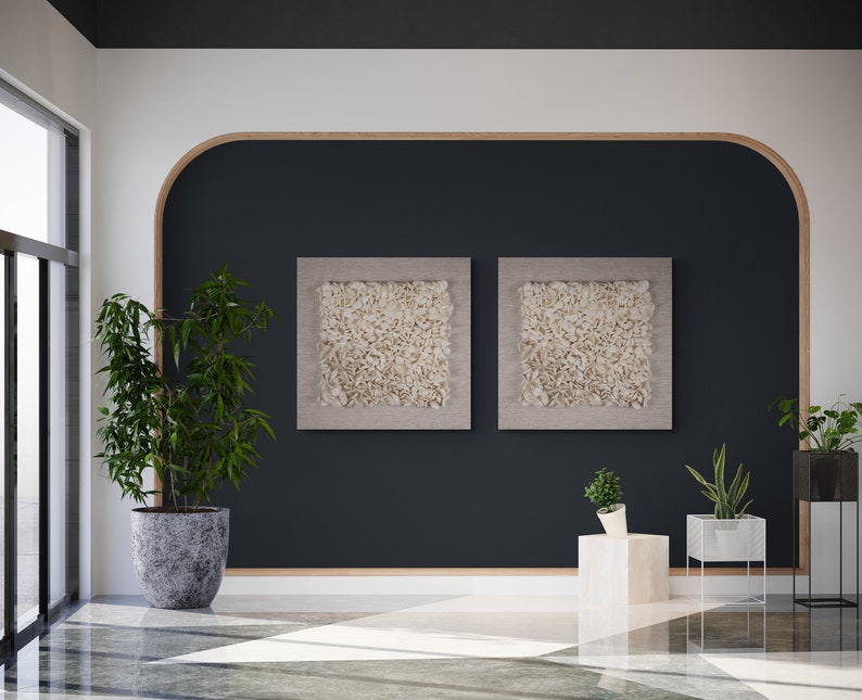 May include: Two framed abstract art prints with a white textured design, hanging on a dark blue wall with a curved wooden frame. The wall is in a modern living room with a potted plant and a side table.